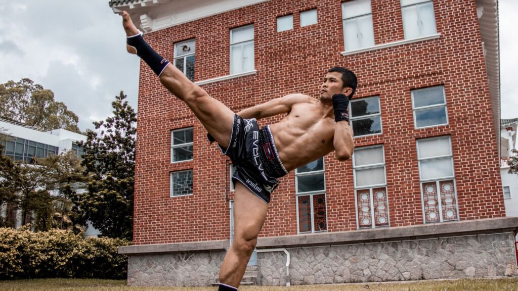 Nong-O throws a Muay Thai head kick in front of a brick building.